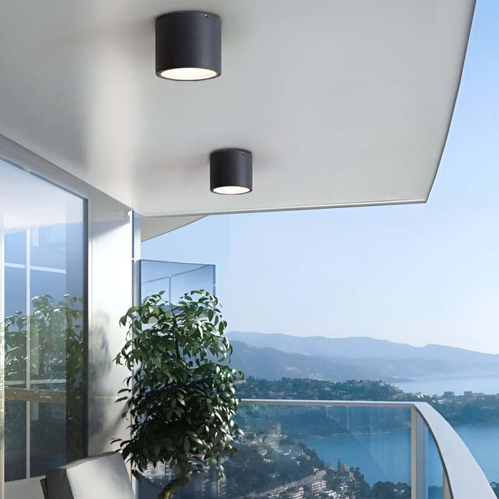 Two black cylindrical outdoor downlights mounted under a balcony overhang with a sea and mountain view.