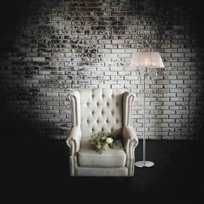 Crystal-accent floor lamp with white shade beside a tufted cream armchair against a distressed white brick wall.