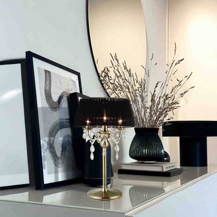 Black-shaded table lamp with crystal drops on a glossy console beside framed art and a black vase.