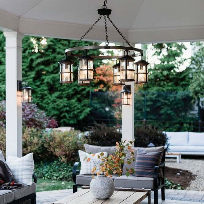 Lantern-style chandelier hanging in a covered outdoor pavilion above a wooden table and cushioned seating, with wall-mounted lanterns and lush garden greenery in the background.