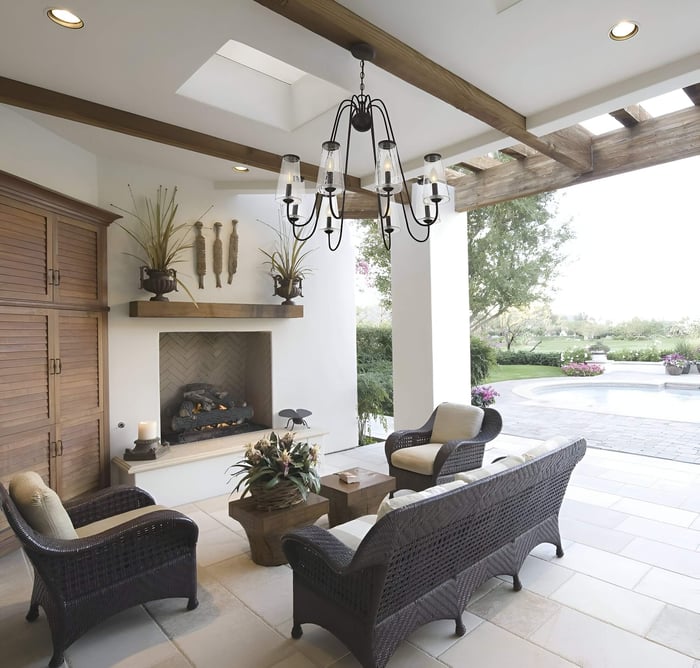 Covered patio seating area with wicker furniture around a fireplace, a black multi-arm chandelier under wood beams, and a view of the garden and pool outside.