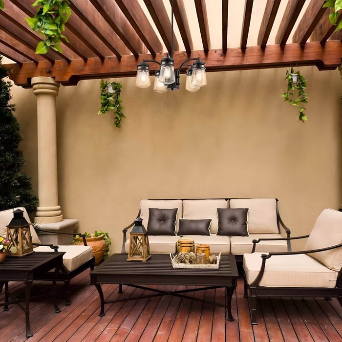 Black outdoor chandelier with five glass shades hanging under a wooden pergola in a neutral-toned patio seating area.
