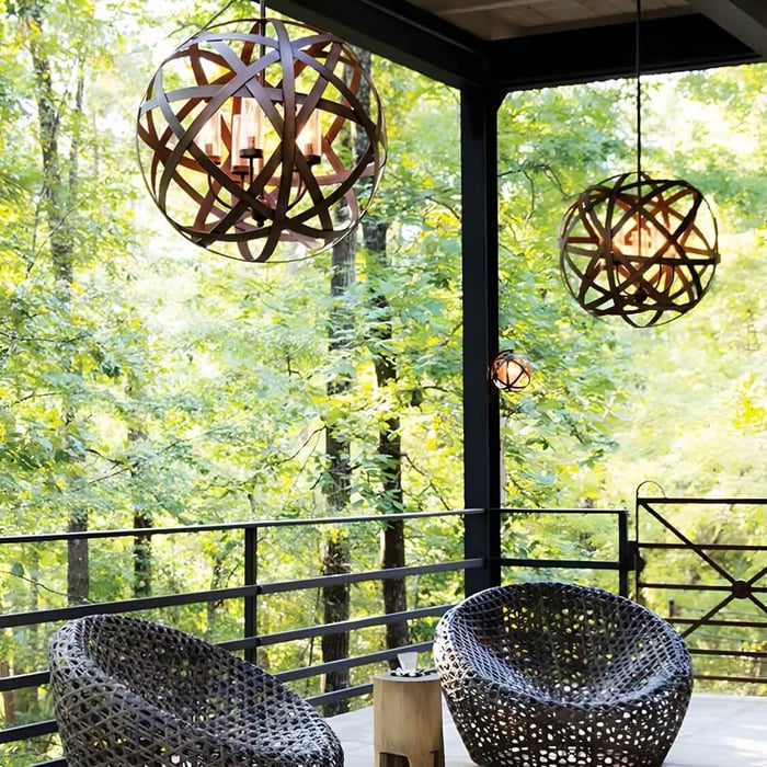 Warm-lit orb-shaped wooden chandeliers hang over a modern forest-view deck with two round woven lounge chairs and a small side table, framed by black railings and posts.