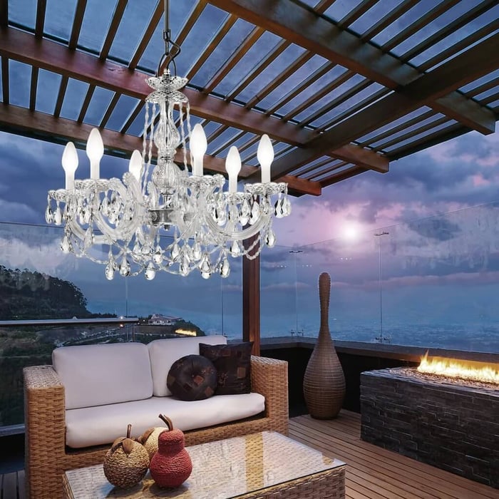 Crystal chandelier hanging under a pergola on a modern outdoor terrace with wicker seating, glass railing, a long fire feature, and a dusk sky over mountains and water.