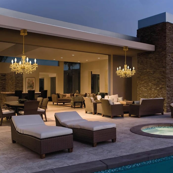 Modern outdoor patio at dusk with wicker lounge chairs, seating area, and two gold chandeliers beside a pool and spa.