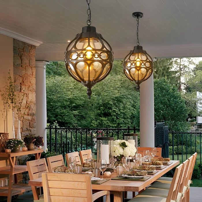 Two ornate globe-shaped pendant chandeliers glow warmly above a long wooden outdoor dining table set for a meal on a covered patio overlooking a lush garden.