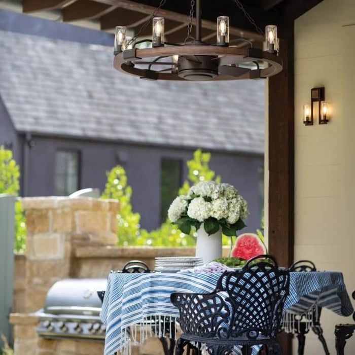 Rustic wagon-wheel chandelier with glass lantern bulbs hanging over an outdoor patio table with a blue striped cloth, hydrangeas, and sliced watermelon.