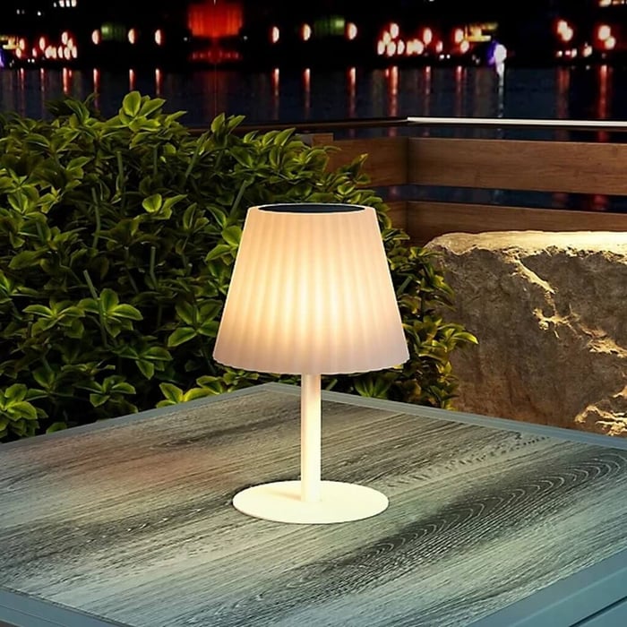 Close-up of a white pleated-shade Outdoor Table Lamps glowing on a gray patio table, with greenery and a stone wall in the background at night.