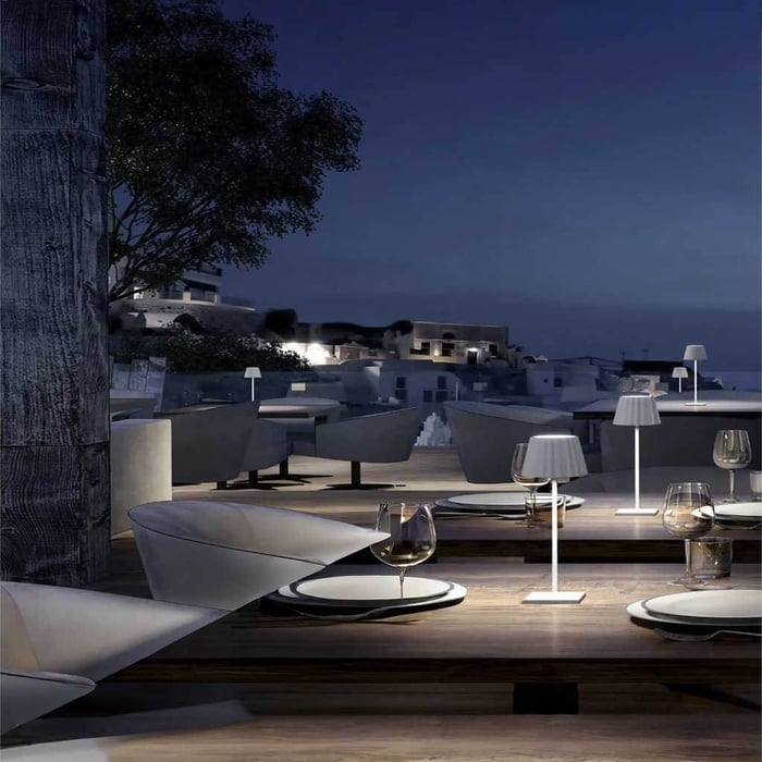 Rooftop terrace at dusk with multiple white cordless Outdoor Table Lamps lighting wooden dining tables against a blue night sky and cityscape backdrop.