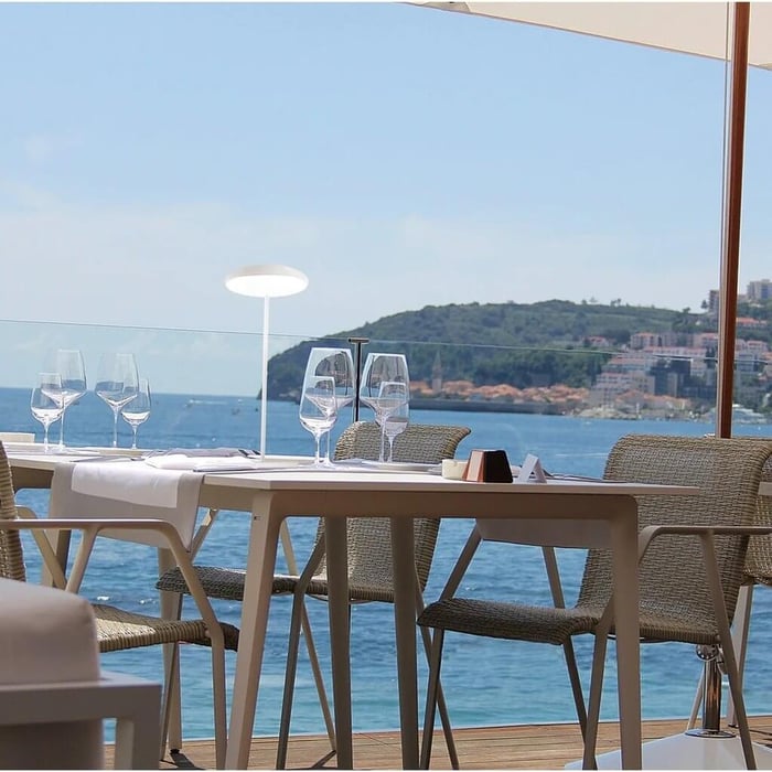 Seaside restaurant table set with glassware and wicker chairs, featuring a slim white cordless Outdoor Table Lamps glowing against a blue ocean and hillside town backdrop on a sunny day.