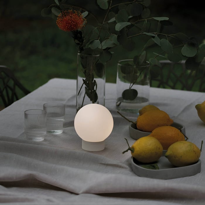 Small spherical white portable lamp on a linen-covered table with lemons, glassware, and green foliage in the background.