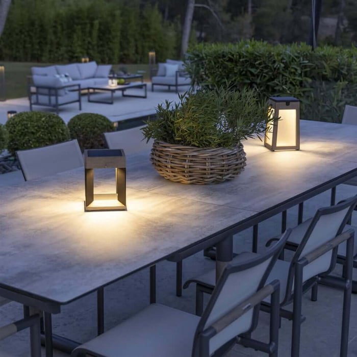 Backyard dining setup with portable lantern-style Outdoor Table Lamps casting warm pools of light on a long stone table, surrounded by chairs and greenery.