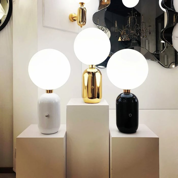 Three glowing globe table lamps on white display plinths white, gold, and black bases in a bright showroom with a sculptural mirror and gold wall sconce behind them.