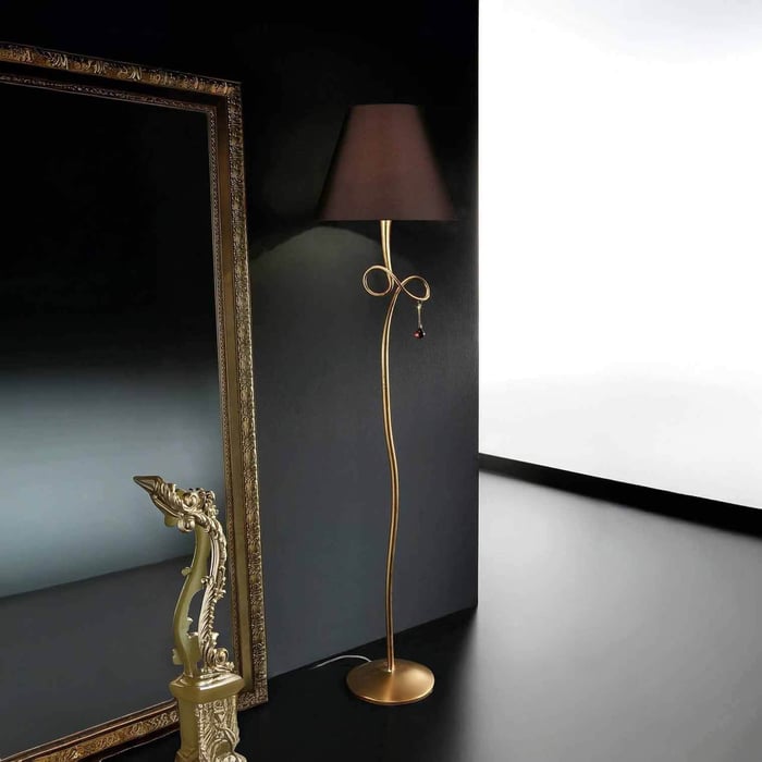 Elegant gold floor lamp with black shade and bow detail beside an ornate framed mirror in a dark room.