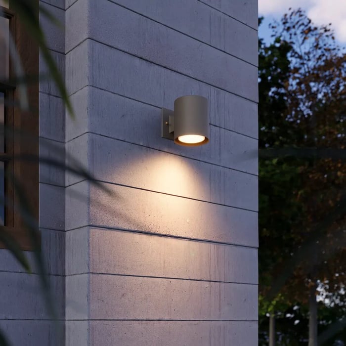 Grey cylindrical wall light projecting a warm pool of light down a textured exterior wall.
