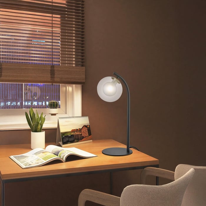Cozy workspace with a wooden desk, modern lamp, open magazine, and potted plant by a window with blinds.