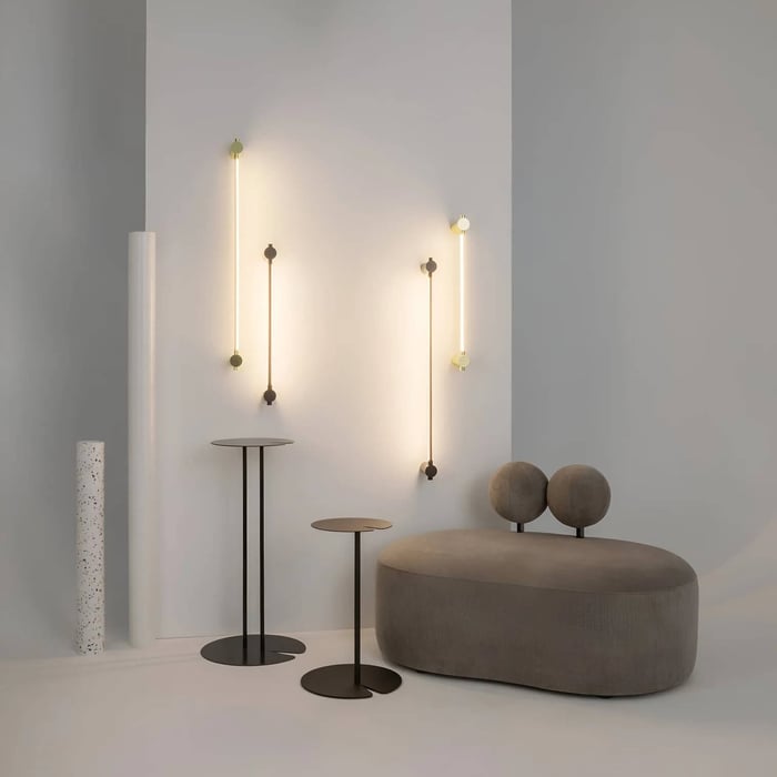 Vertical LED wall lights with round brass and bronze mounts above a sculptural pebble sofa and slim pedestal tables.