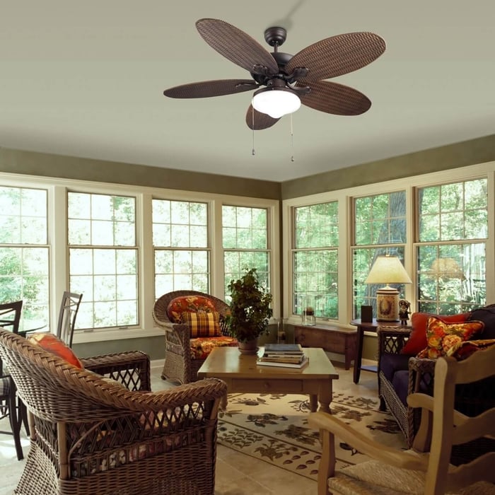 Tropical-style ceiling fan with wicker blades and light in a sunroom with natural views.