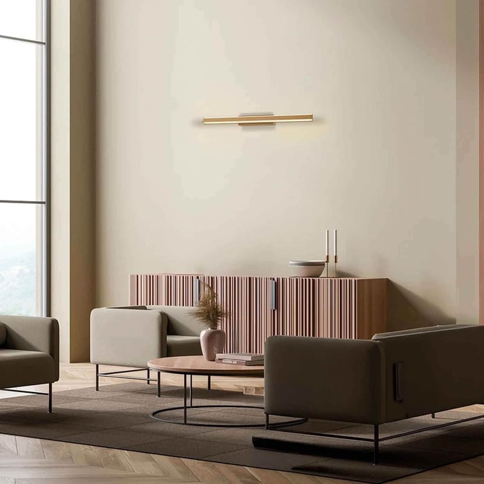Minimalist living room with warm beige walls, a slim brushed-brass LED picture light above a fluted wood credenza, low neutral sofas, round coffee table, and daylight from a tall window.