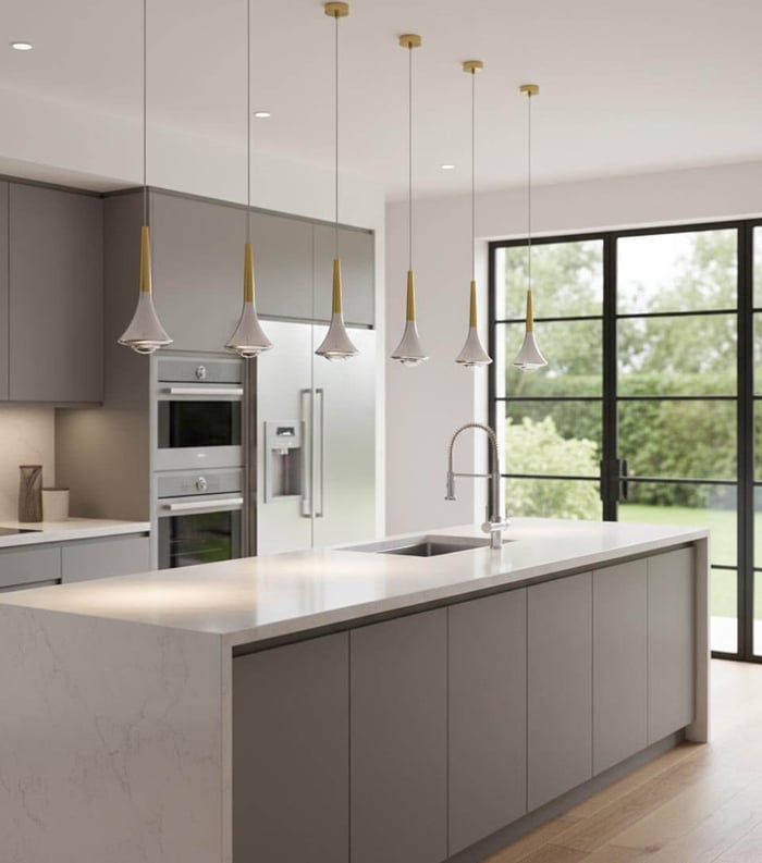 Bright modern kitchen with a long white island, gray cabinets, stainless steel appliances, and a row of slim white-and-gold pendant lights hanging above, next to large black-framed glass doors overlooking a green yard.