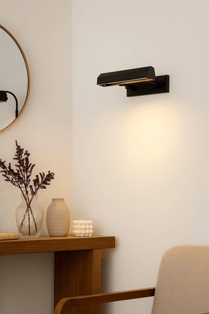 Matte black picture light on a white wall illuminating above a wooden console with minimalist vases, a round mirror edge, and a beige chair.