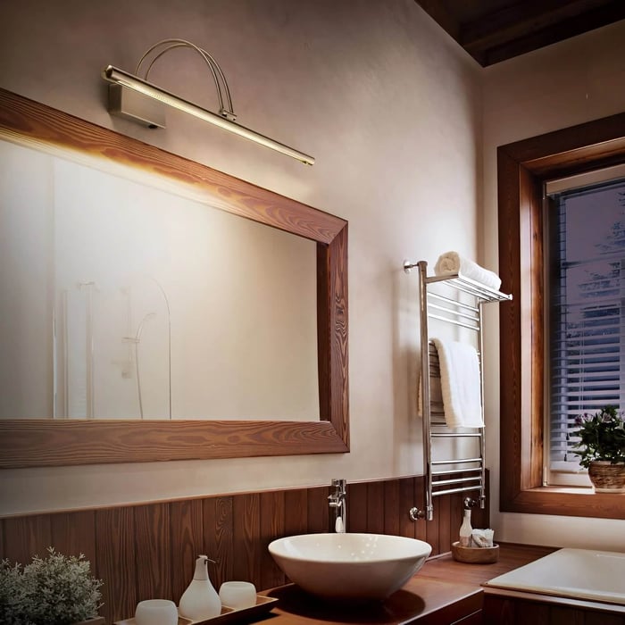 Warm, wood-accented bathroom with a brushed-nickel arched LED picture light above a large wood-framed mirror, vessel sink on a wooden vanity, chrome towel warmer, and window with blinds.