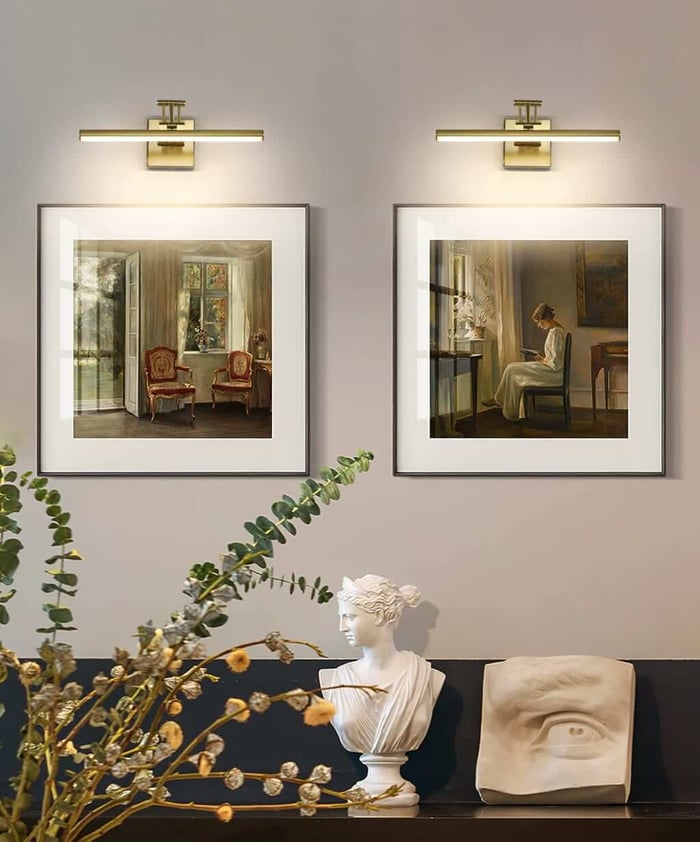Two framed paintings lit by slim brass picture lights above a console with a classical bust, sculpted eye fragment, and dried greenery against a taupe wall.