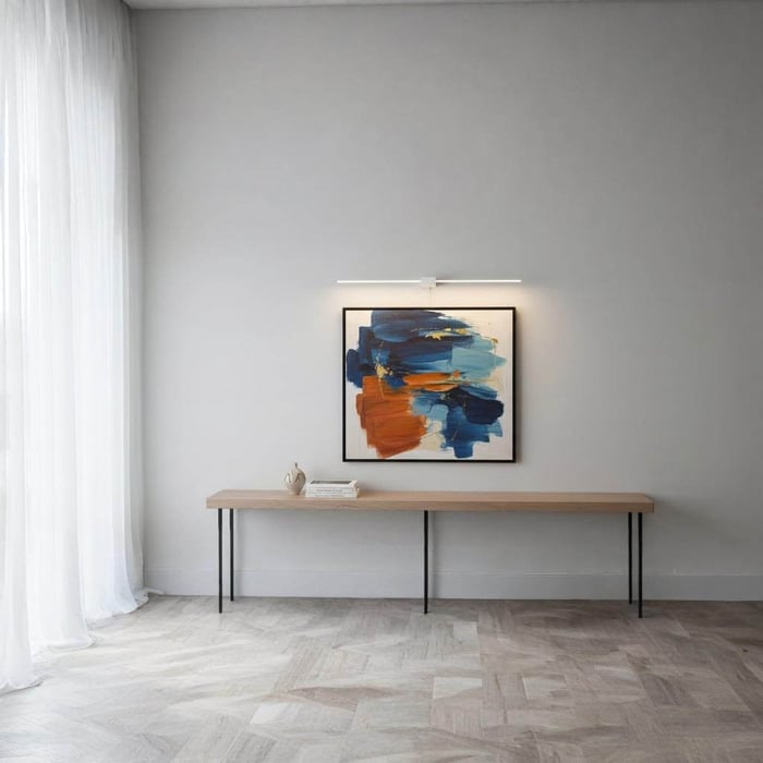 Slim white picture light illuminating a blue-and-orange abstract painting above a long wood console against a light gray wall with sheer curtains.