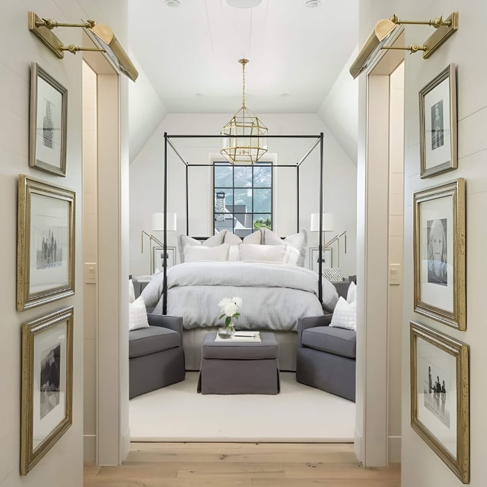 A refined bedroom entrance view with gold picture lights illuminating framed art on both sides of the doorway. The bedroom features a canopy bed, grey chairs, and a gold geometric chandelier.