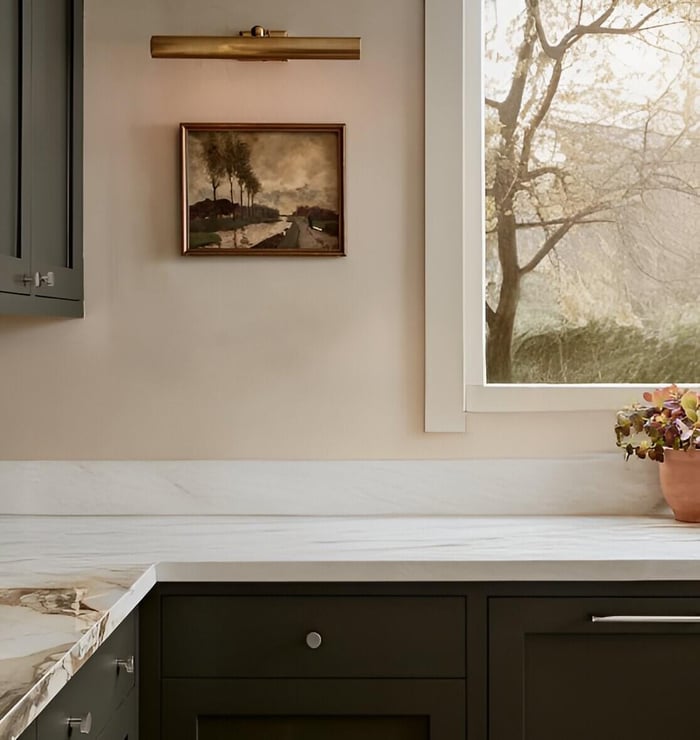 Brass picture light illuminating a small landscape painting above marble countertops and dark green kitchen cabinets beside a window.