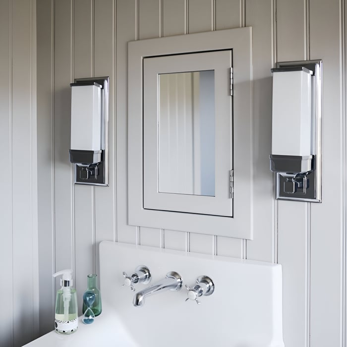 A white bathroom vanity mirror surrounded by vertical wall sconces with chrome finishes, creating a sleek, modern look. The setup is centered above a white sink with silver faucets, adding a clean, sophisticated touch.