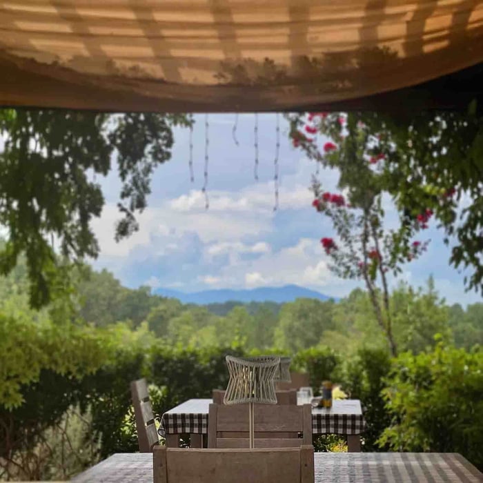 Outdoor dining area under a canopy with a table lamp as the centrepiece, overlooking a scenic garden and distant hills.