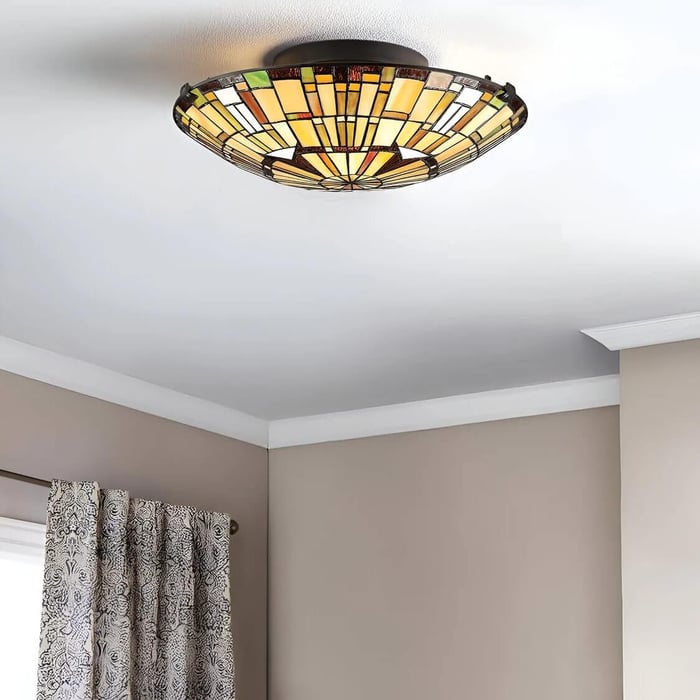 Stained-glass flush-mount ceiling light with warm yellow and amber panels, installed on a white ceiling above beige walls and patterned curtains.