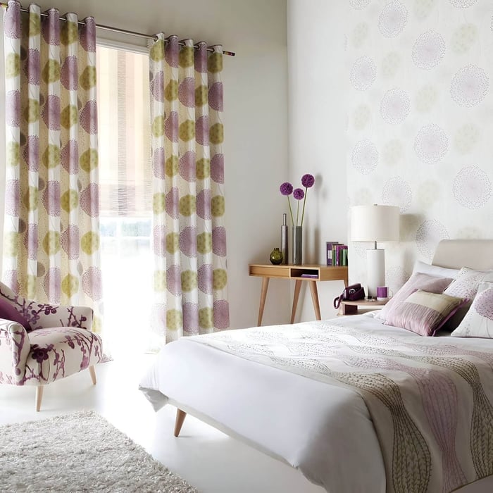 Playful pastel bedroom with white and lavender bedding, floral curtains, and ceramic table lamp on a wooden desk.