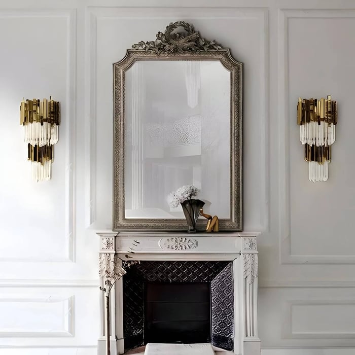 Elegant interior with a decorative white fireplace, a large ornate mirror above it, and two gold crystal wall sconces on each side, creating a luxurious and sophisticated ambiance.