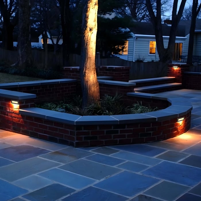 Curved red brick seating walls with rectangular wall lights illuminating a tree-centered patio at night.