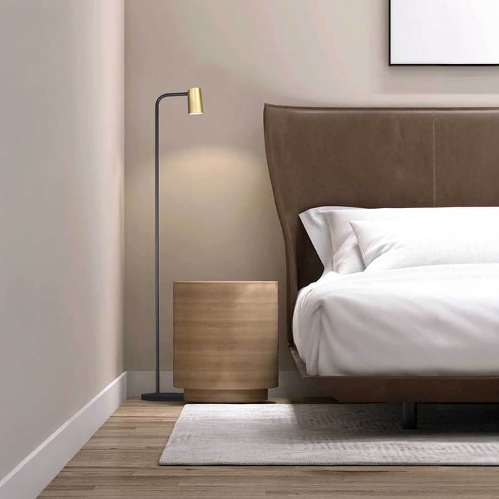 Minimal black-and-brass industrial floor lamp beside a bed and wooden nightstand in a calm modern bedroom.