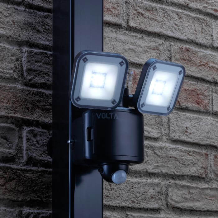 Black dual-head LED security floodlight with a motion sensor mounted on a brick exterior wall, both lights shining brightly at night.