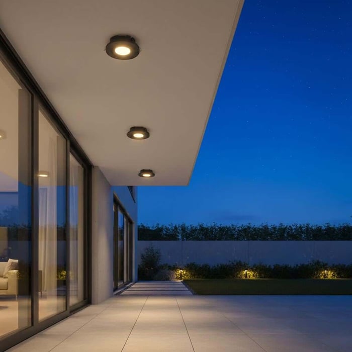 Modern patio at night with recessed ceiling downlights above a tiled terrace, glass walls on the left, and a deep blue evening sky over a landscaped yard.