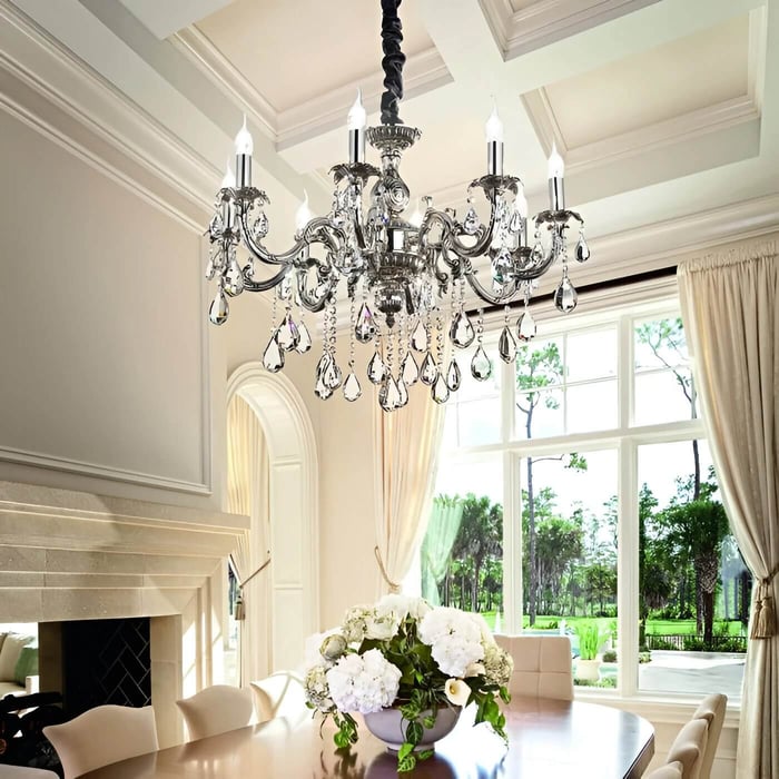 Classic chrome-and-crystal chandelier with candle-style bulbs hanging above a dining table in a bright room with large windows and soft curtains.