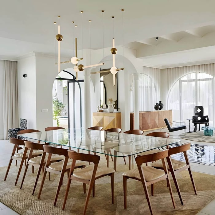 Gold and white linear pendant cluster above a glass dining table in a bright, modern open-plan room with arched doorways.