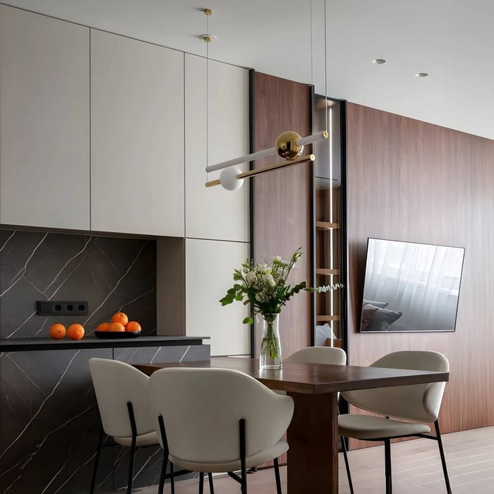 A contemporary dining area featuring a minimalist gold and white linear pendant light hanging over a dark wood table. The background shows a modern kitchen with dark marble backsplashes and a wood-paneled wall with an integrated television.