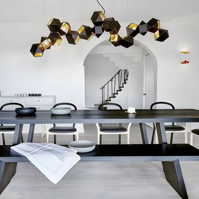 Sculptural black-and-gold geometric linear pendant light spanning above a long dining table in a minimalist white dining room.