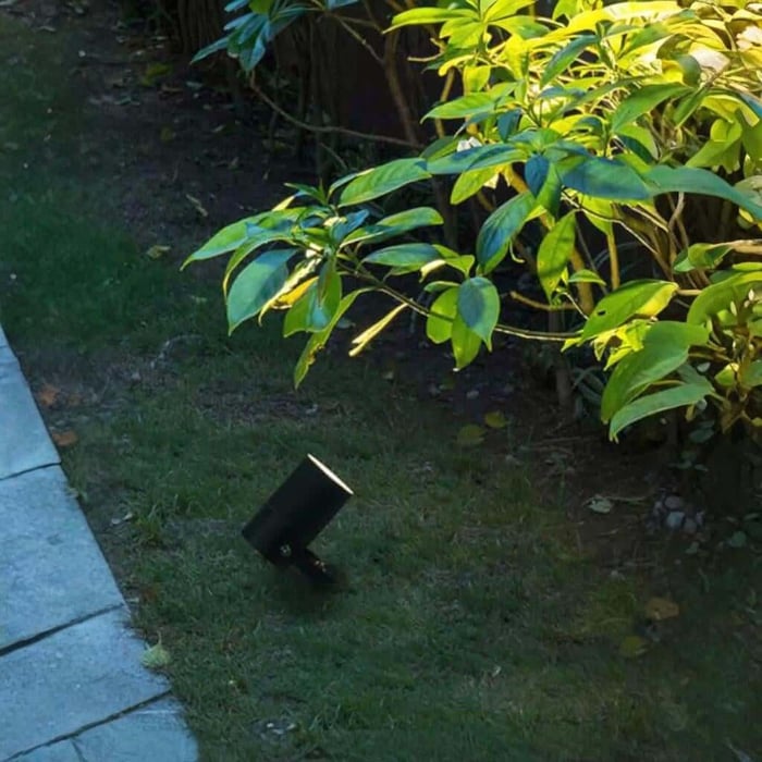 Black garden spike spotlight shining warm light onto shrubs beside a patio edge.