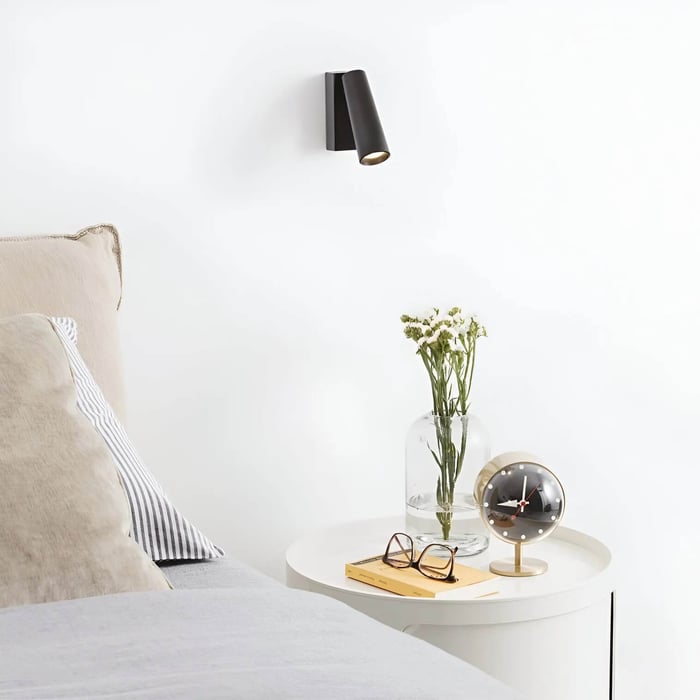 Bright white bedroom scene featuring a small black wall-mounted spotlight over a round bedside table with flowers and décor.