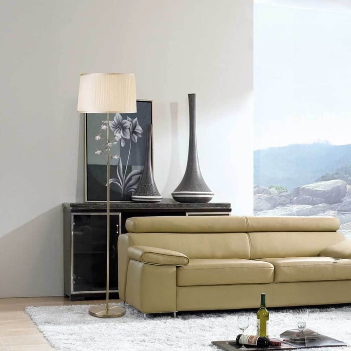 Modern living room with a beige leather sofa, slim brass floor lamp with a pleated shade, black sideboard with tall vases, and a large window.