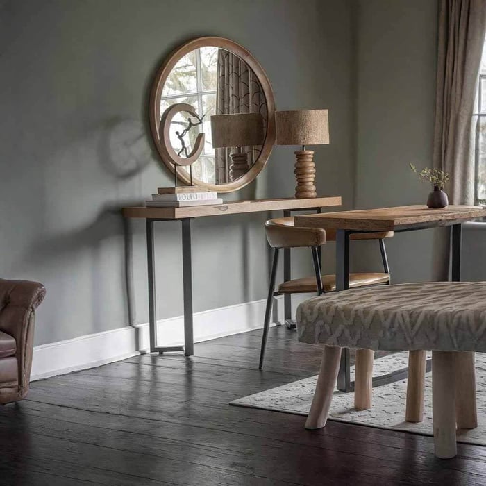 A rustic console table with a round wooden mirror, sculptural wooden table lamp, leather chair, and upholstered bench in a soft gray, modern farmhouse-style room.