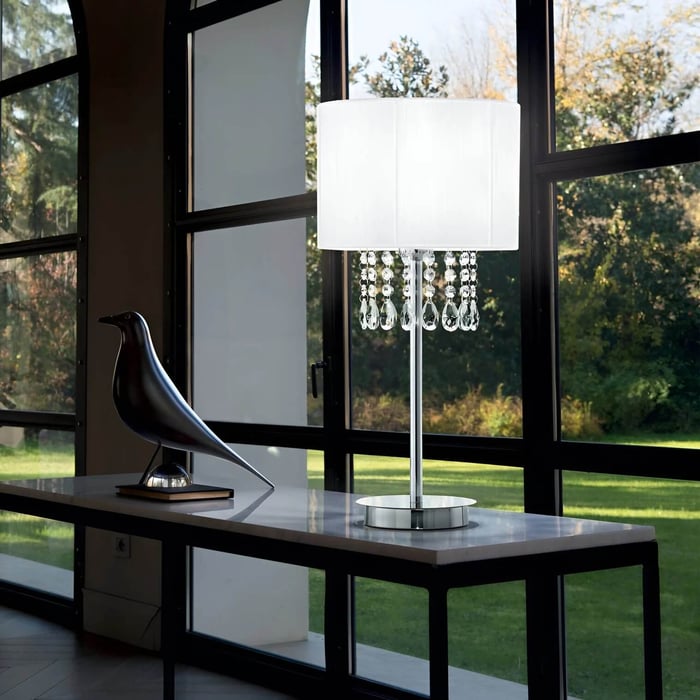 A modern crystal table lamp with a white drum shade and hanging glass droplets sits on a sleek console table in front of large black-framed windows overlooking a green garden.