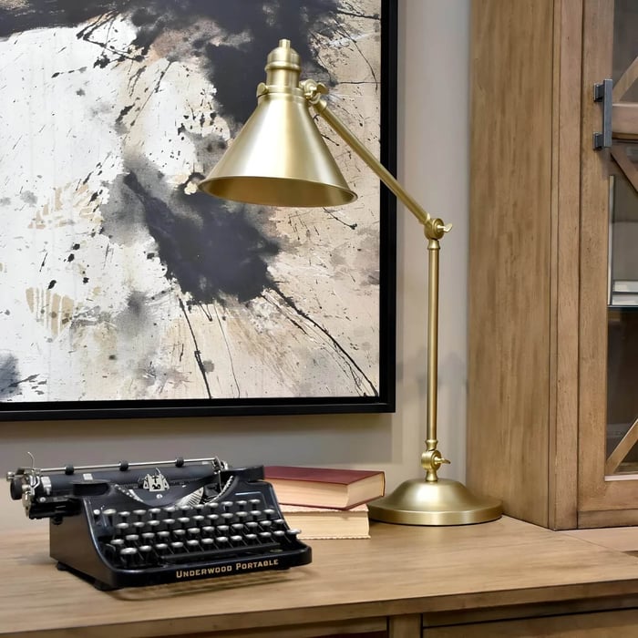 A brass adjustable desk lamp stands on a wooden cabinet beside a vintage Underwood typewriter and books, set against a large abstract black-and-white painting.