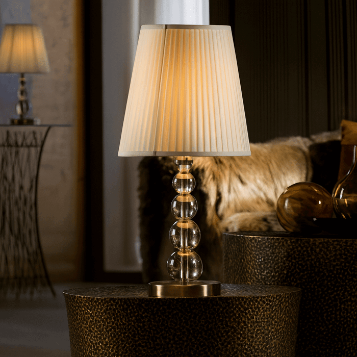 A warm, ambient table lamp with a pleated beige lampshade and a stacked glass orb base sits on a textured bronze side table in a cozy, softly lit living room.